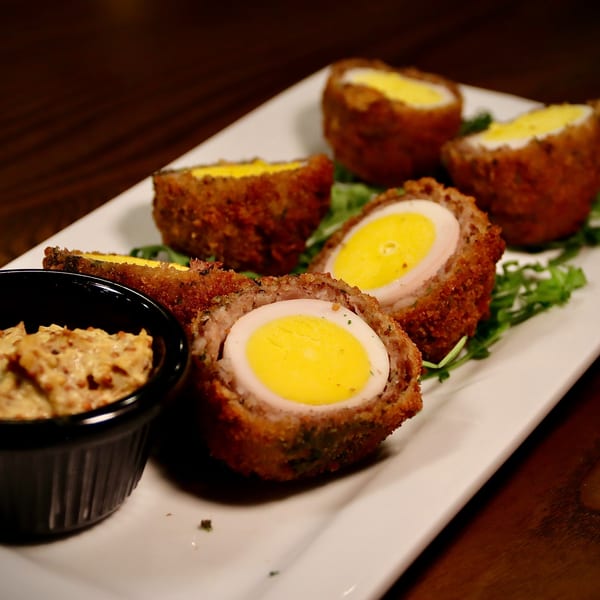 Several golden-brown Scotch eggs, halved to show bright yellow yolks, served with a dipping sauce.