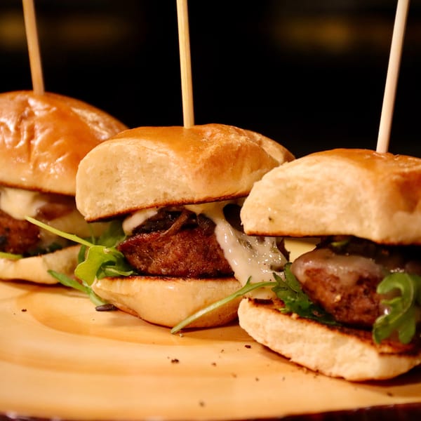 Three American sliders with meat, cheese, and arugula on a wooden board.