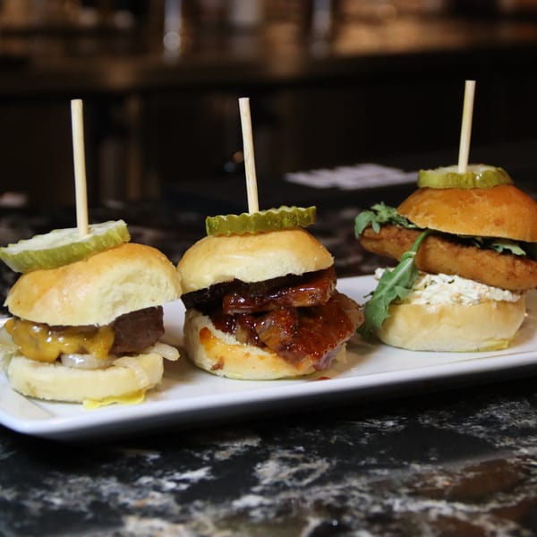 Three gourmet sliders, including cheeseburger, BBQ, and fried fish, on a white plate.