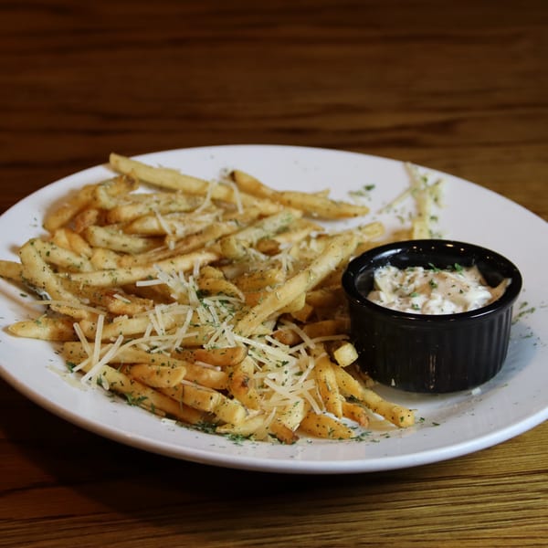 A white plate of golden french fries topped with shredded cheese and herbs, with a side of creamy dip.