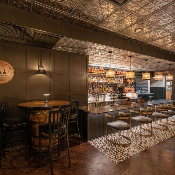 The Barrel Room at Beckett’s bar area with a barrel table, bar stools, and liquor bottles.